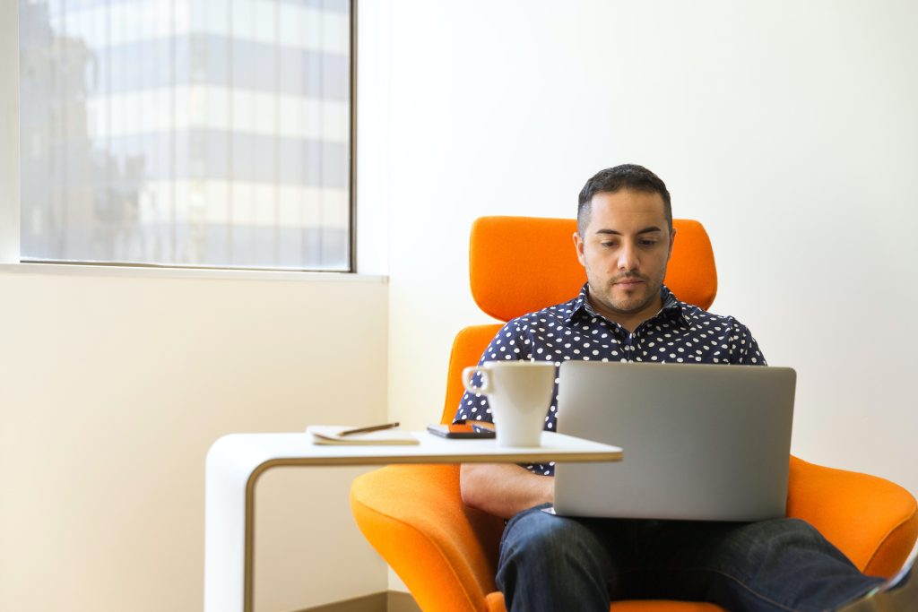 Man sitting on an orange chair in a brightly light office. He is typing on his laptop which is on the table in front of him.