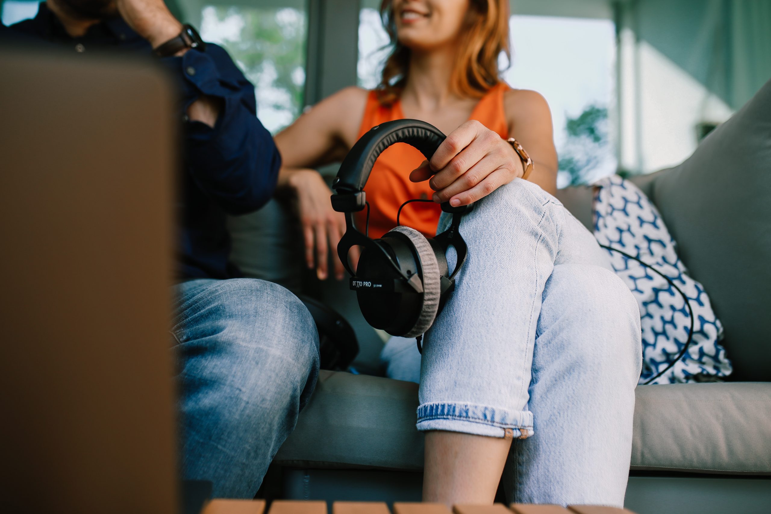 Two people sitting down. The woman is wearing an orange top, jeans and is holding a set of headphones.