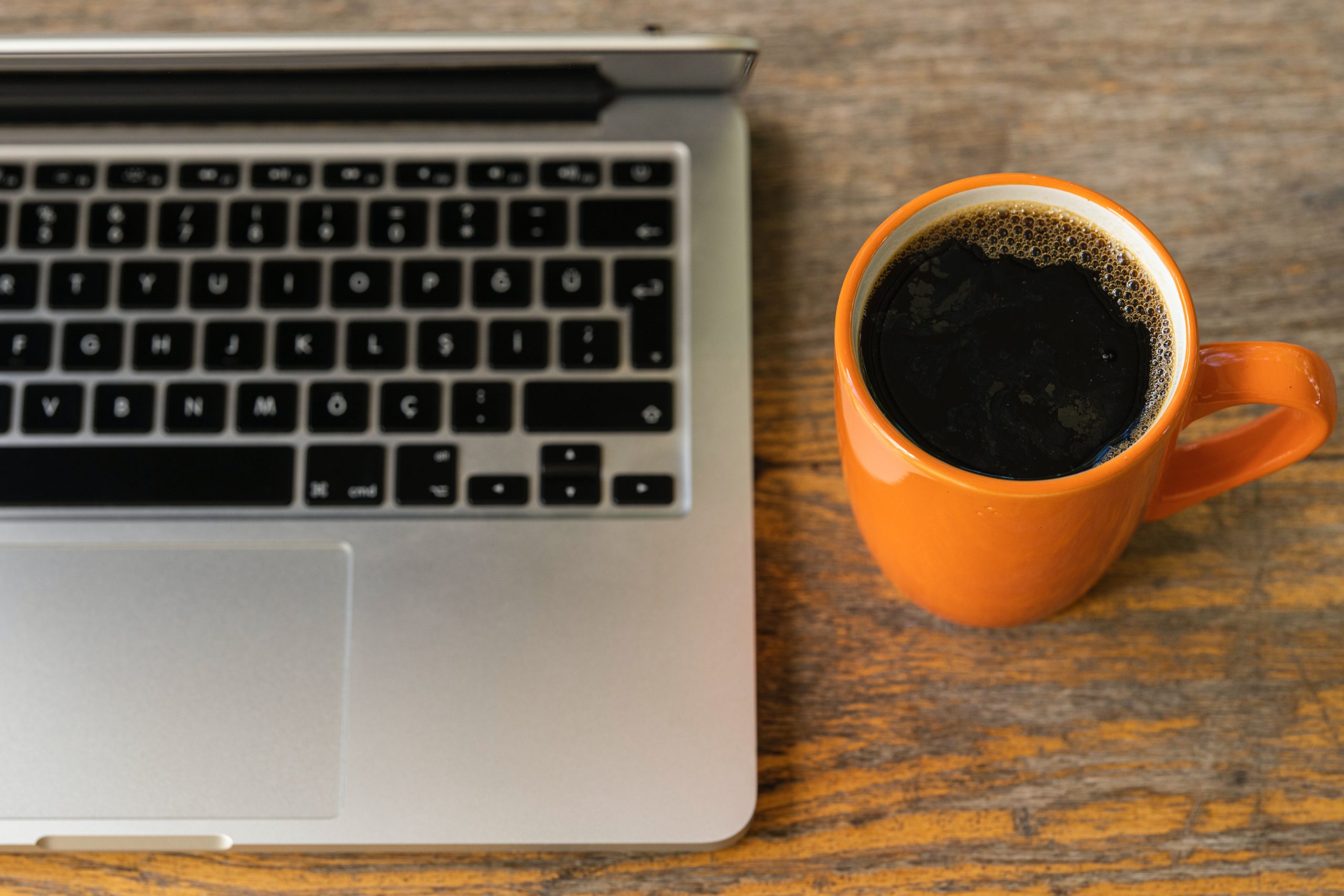 A orange mug of coffee sit next to a silver, opened laptop on a wooden desk.