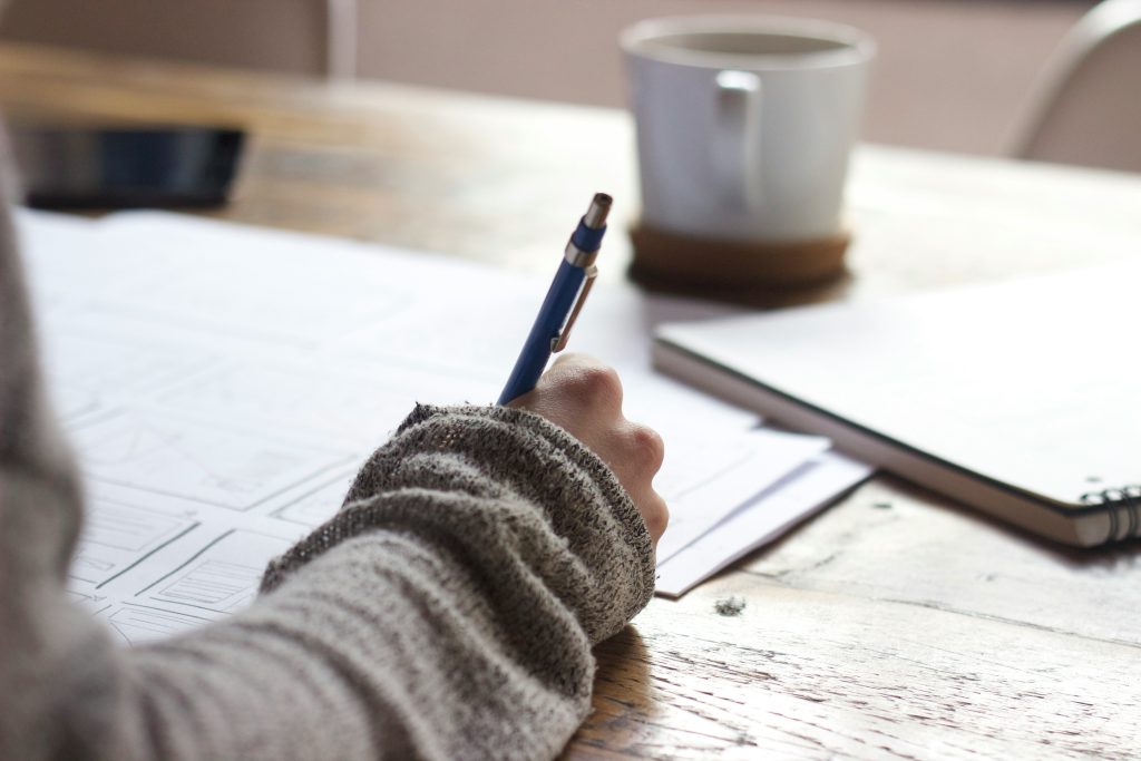 person writing in a notebook which is on a wooden desk. There is a mug with a hot drink in the background.