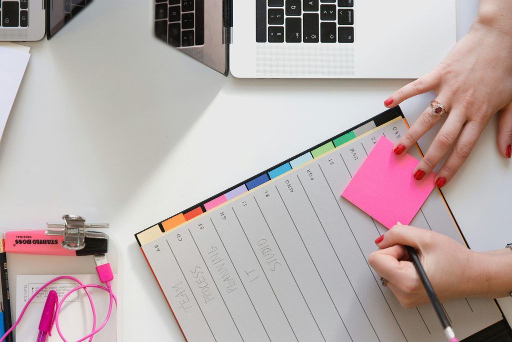 a women is writing on a pink post it note which is on a desk. There is a laptop to her right and a highlighter and headhphones also on the desk.