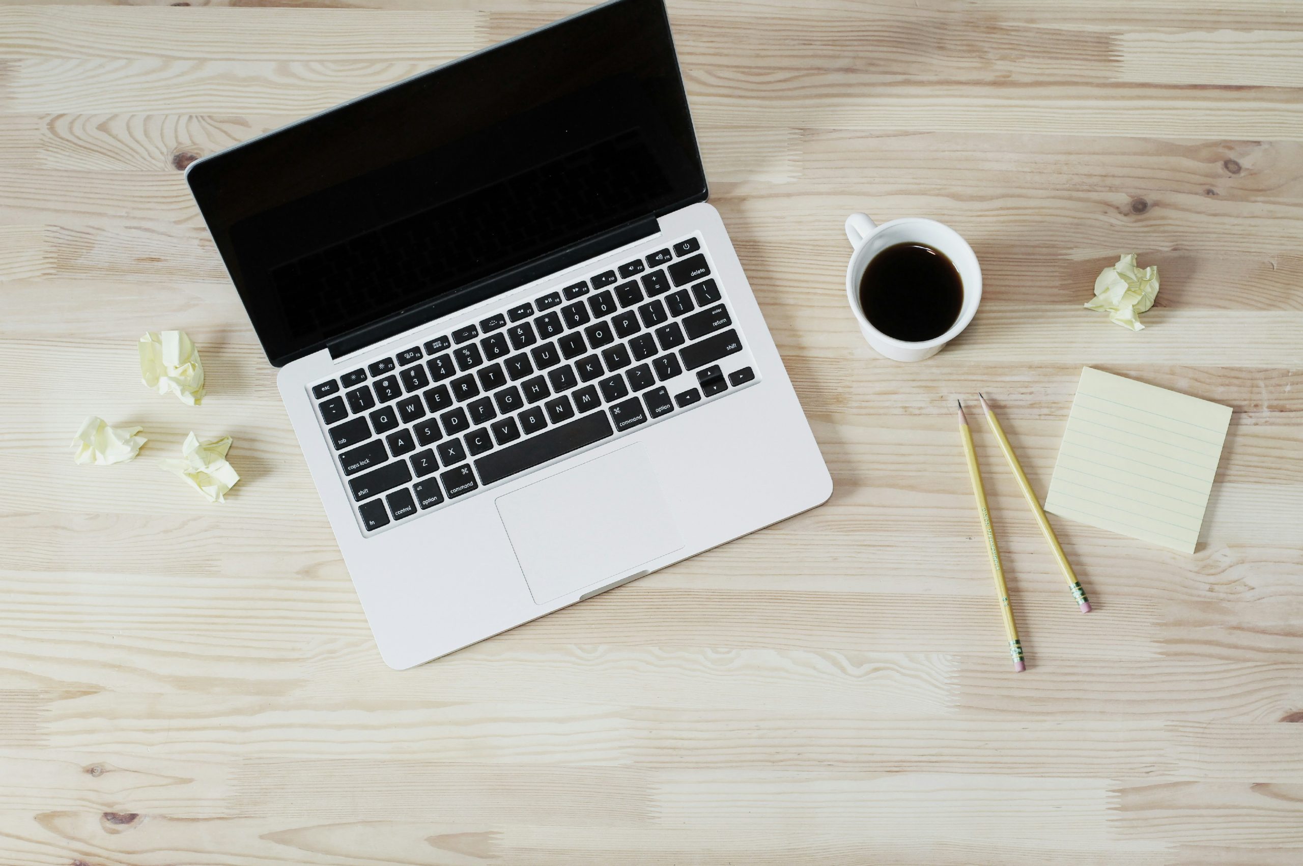 a laptop is open on a wooden desk. There is a cup of coffee next to it, along with pencils and scrunched up papers.