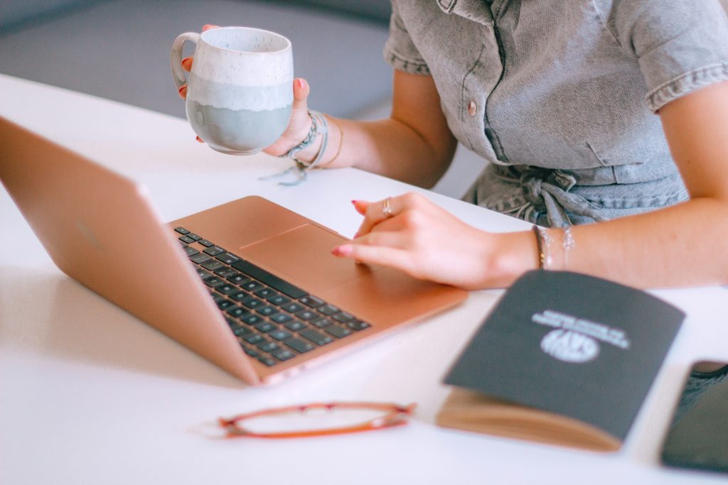 woman at a desk using her laptop. She is drinking a hot drink and her notebook and glasses are also on the desk.