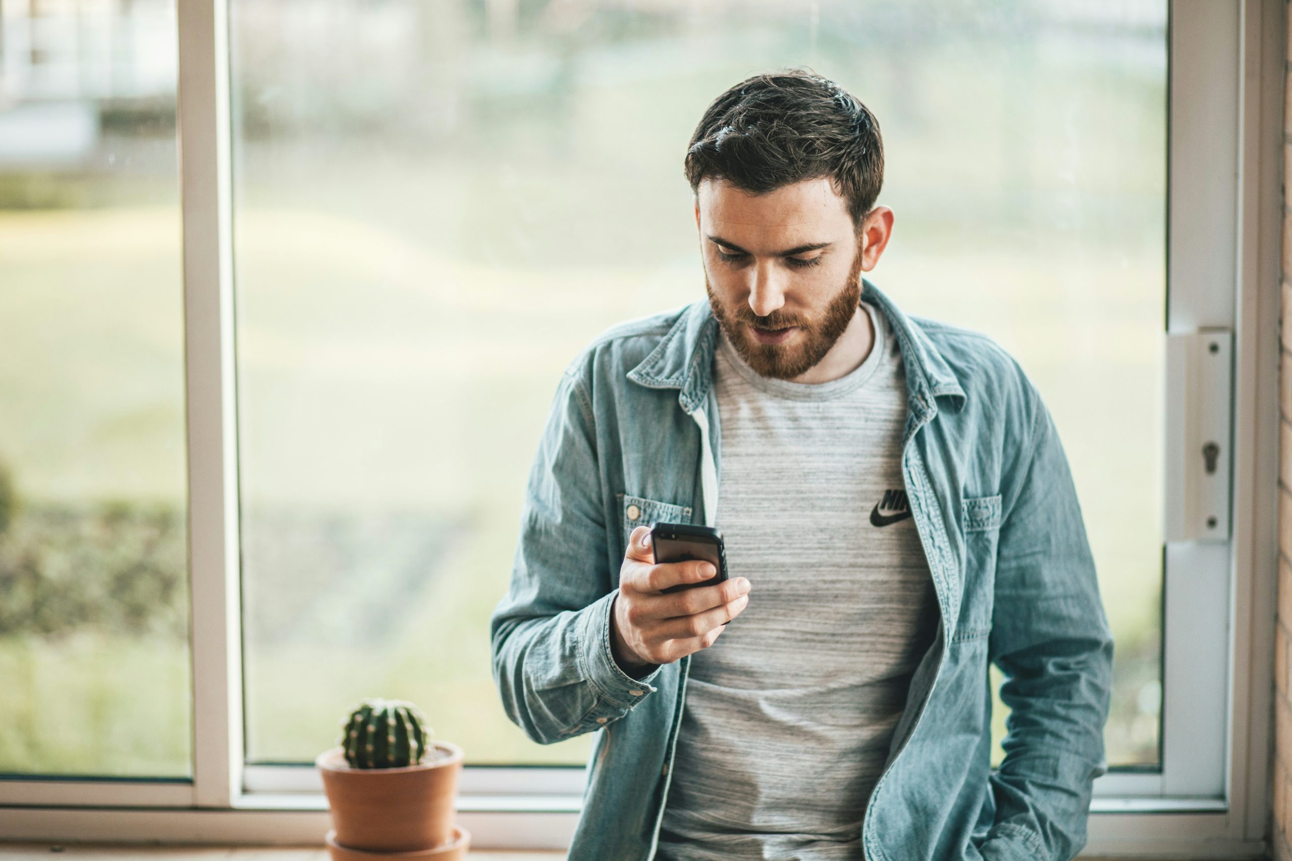 man in a denim shirt is sitting on a window ledge, looking at his phone.